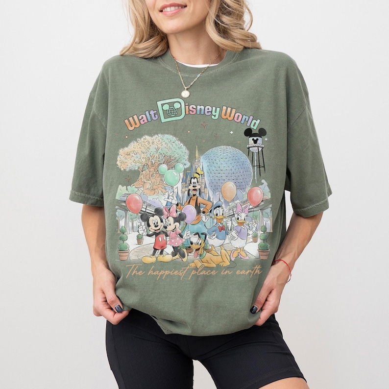 May include: Olive green t-shirt featuring a colorful graphic of Disney characters and landmarks, including Mickey Mouse, Minnie Mouse, and the Spaceship Earth. The text "Walt Disney World" and "The happiest place in earth" are also displayed.