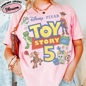 Puede incluir: Una camiseta de manga corta de color rosa claro con un estampado gr&aacute;fico que incluye el texto Disney Pixar Toy Story 5. El dise&ntilde;o presenta ilustraciones de Buzz Lightyear, Woody, Rex, Jessie sobre Perdig&oacute;n y otros personajes.