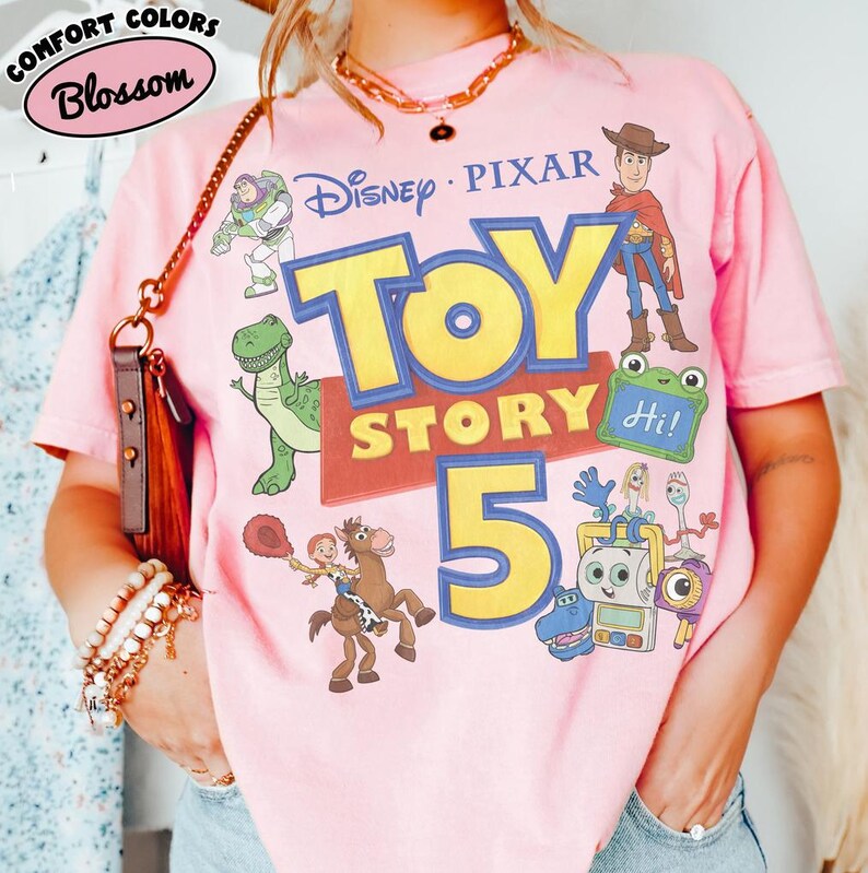 Puede incluir: Una camiseta de manga corta de color rosa claro con un estampado gr&aacute;fico que incluye el texto Disney Pixar Toy Story 5. El dise&ntilde;o presenta ilustraciones de Buzz Lightyear, Woody, Rex, Jessie sobre Perdig&oacute;n y otros personajes.