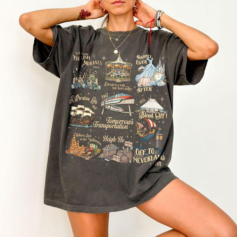 K&ouml;nnte beinhalten: Dunkelgraues T-Shirt mit Themenpark-Bildern und S&auml;tzen wie &bdquo;Foolish Mortals&ldquo; und &bdquo;Happily Ever After&ldquo;. Das Design umfasst ein Karussell, ein Piratenschiff und eine Einschienenbahn. Der Text &bdquo;Tomorrow's Transportation&ldquo; ist ebenfalls sichtbar.