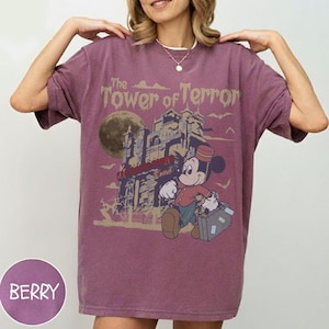 Puede incluir: Camiseta de color bayas con un gr&aacute;fico de Mickey Mouse como botones frente a un hotel embrujado, con el texto "The Tower of Terror". El dise&ntilde;o incluye una luna llena y p&aacute;jaros volando.