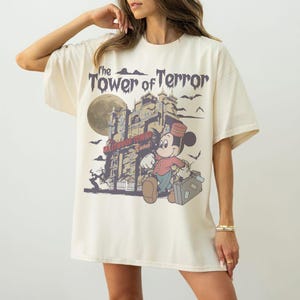 Puede incluir: Camiseta color crema con un dise&ntilde;o de Mickey Mouse como botones frente a un edificio embrujado. El texto dice "The Tower of Terror" y "Hollywood Tower Hotel". El dise&ntilde;o incluye una luna llena y murci&eacute;lagos.