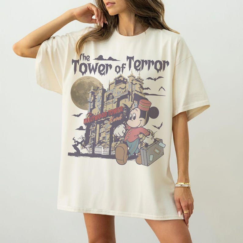 Puede incluir: Camiseta color crema con un dise&ntilde;o de Mickey Mouse como botones frente a un edificio embrujado. El texto dice "The Tower of Terror" y "Hollywood Tower Hotel". El dise&ntilde;o incluye una luna llena y murci&eacute;lagos.