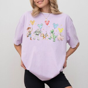 May include: A light purple t-shirt featuring a colorful graphic of cartoon characters holding Mickey Mouse-shaped balloons. The characters include a cowboy, an astronaut, a dog, a dinosaur, and a pig. The design is whimsical and playful.