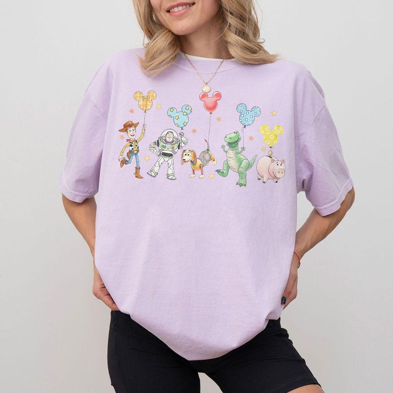 May include: A light purple t-shirt featuring a colorful graphic of cartoon characters holding Mickey Mouse-shaped balloons. The characters include a cowboy, an astronaut, a dog, a dinosaur, and a pig. The design is whimsical and playful.