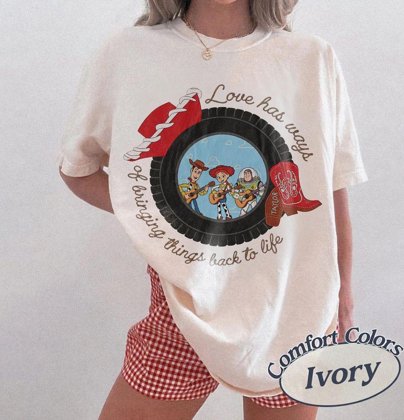 May include: An ivory t-shirt featuring a graphic of a black tire framing a scene with three cartoon characters playing guitars. A red cowboy hat and a red cowboy boot are positioned on the tire. The text reads, Love has ways of bringing things back to life.
