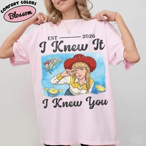 May include: A light pink t-shirt featuring a graphic print of a blonde character wearing a red cowboy hat, surrounded by small cartoon figures. The text reads EST 2026, I Knew It, and I Knew You in black lettering. The shirt has a crew neckline.