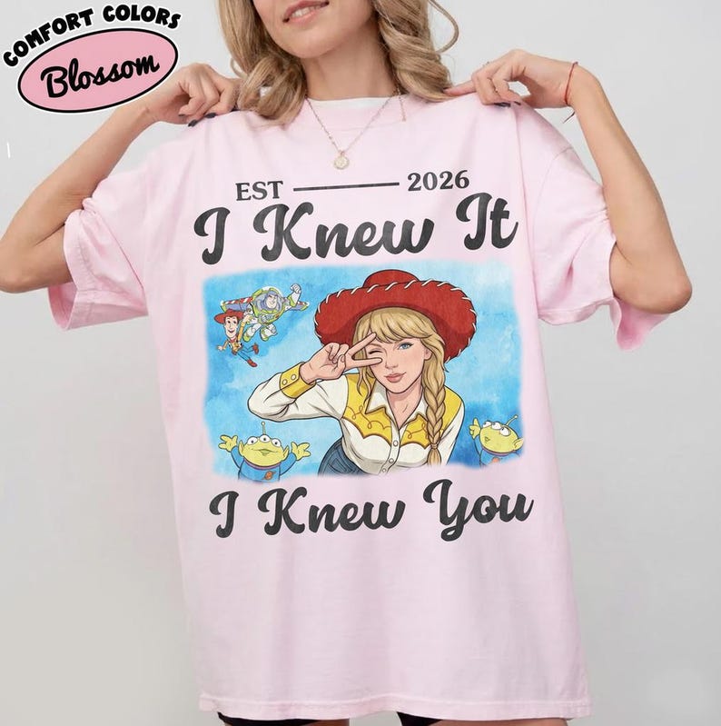 May include: A light pink t-shirt featuring a graphic print of a blonde character wearing a red cowboy hat, surrounded by small cartoon figures. The text reads EST 2026, I Knew It, and I Knew You in black lettering. The shirt has a crew neckline.