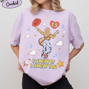 Puede incluir: Una camiseta de color morado claro con un gr&aacute;fico de una persona de cabello rubio que viste una camisa de cuadros amarilla y pantalones con estampado de vaca, sosteniendo una guitarra y un sombrero rojo. El dise&ntilde;o incluye un coraz&oacute;n rojo con "TS", nubes, estrellas, un cohete y el texto "I KNEW IT I KNEW YOU".