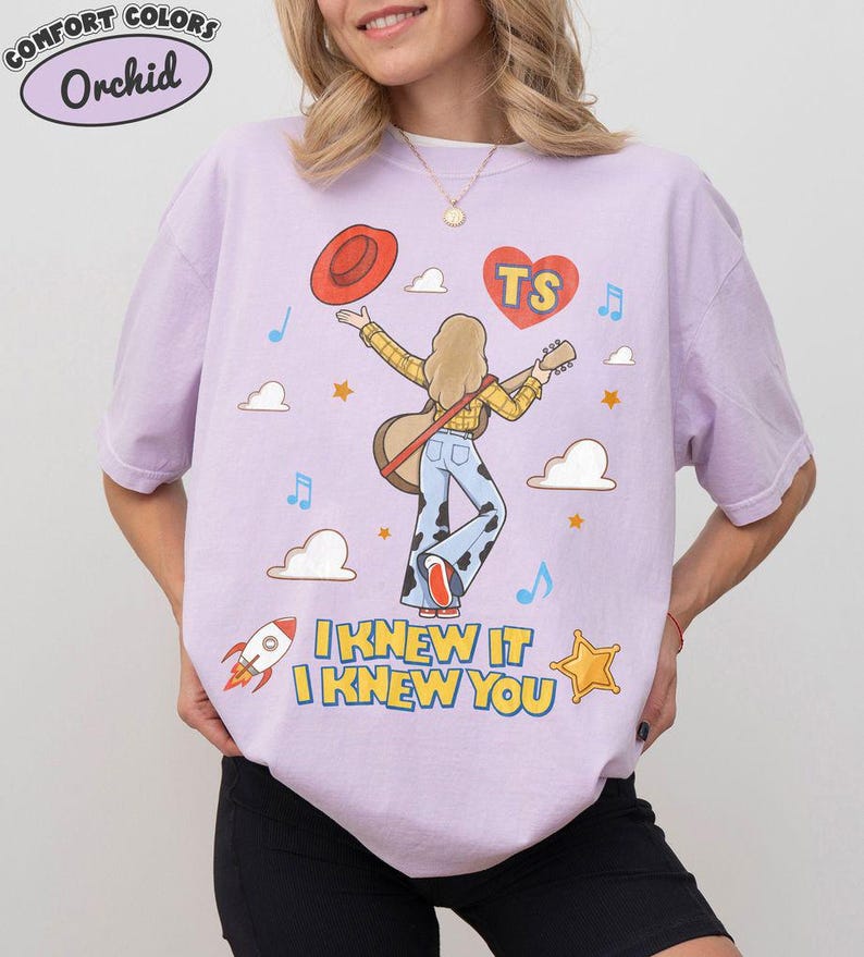 Puede incluir: Una camiseta de color morado claro con un gr&aacute;fico de una persona de cabello rubio que viste una camisa de cuadros amarilla y pantalones con estampado de vaca, sosteniendo una guitarra y un sombrero rojo. El dise&ntilde;o incluye un coraz&oacute;n rojo con "TS", nubes, estrellas, un cohete y el texto "I KNEW IT I KNEW YOU".