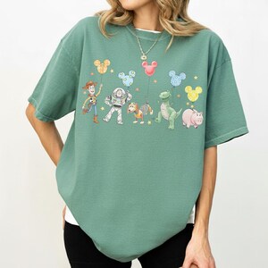 May include: A green t-shirt featuring a colorful graphic of Toy Story characters holding Mickey Mouse-shaped balloons. The design includes Woody, Buzz Lightyear, Rex, Hamm, and Slinky Dog. The shirt has a relaxed fit.