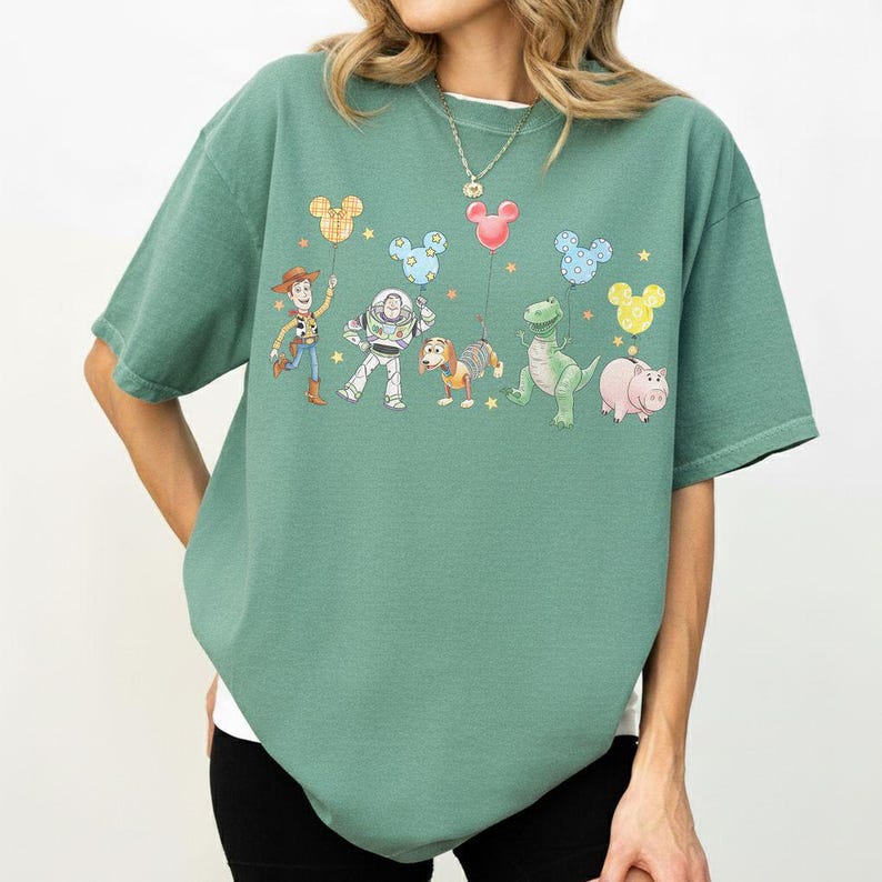 May include: A green t-shirt featuring a colorful graphic of Toy Story characters holding Mickey Mouse-shaped balloons. The design includes Woody, Buzz Lightyear, Rex, Hamm, and Slinky Dog. The shirt has a relaxed fit.