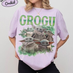 May include: A light purple t-shirt featuring a graphic print of multiple small green creatures with large ears and tan robes. The text GROGU is printed in large green letters above the creatures, which are surrounded by green smoke effects.