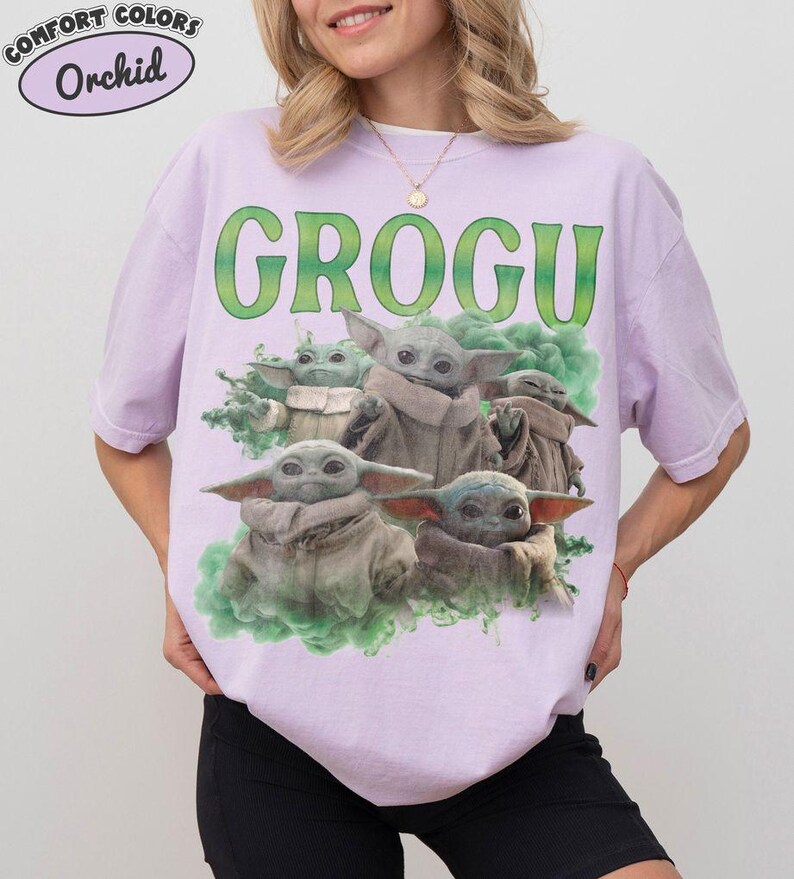 May include: A light purple t-shirt featuring a graphic print of multiple small green creatures with large ears and tan robes. The text GROGU is printed in large green letters above the creatures, which are surrounded by green smoke effects.