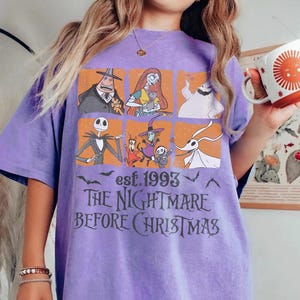 May include: A lilac t-shirt with a graphic from "The Nightmare Before Christmas." The design showcases Jack Skellington, Sally, and other characters in orange squares. The text "est. 1993" and "THE NIGHTMARE BEFORE CHRISTMAS" are printed below the characters.