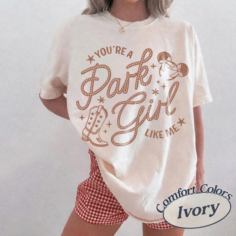 Op de afbeelding: Een ivoorkleurig T-shirt met korte mouwen en bruine grafische tekst met de tekst "YOU'RE A Park Girl LIKE ME" in een touwachtig lettertype. Het ontwerp bevat een cowboylaars, muizenoortjes met strikjes en kleine sterretjes. Het shirt wordt gedragen met een rood-witte gingham short.