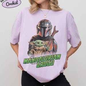 May include: A light purple t-shirt featuring a graphic print of The Mandalorian and Grogu. The Mandalorian wears a metallic helmet and armor, while Grogu is positioned in front. The text "THE MANDALORIAN AND GROGU" appears in green below.