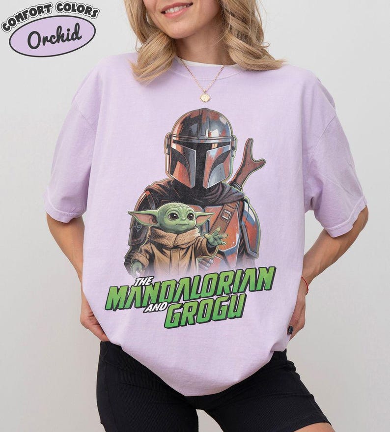 May include: A light purple t-shirt featuring a graphic print of The Mandalorian and Grogu. The Mandalorian wears a metallic helmet and armor, while Grogu is positioned in front. The text "THE MANDALORIAN AND GROGU" appears in green below.