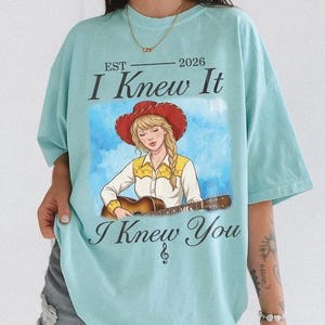 May include: A light blue t-shirt featuring a graphic of a person with a blonde braid wearing a red cowboy hat and a yellow and white shirt while playing an acoustic guitar. The text reads EST 2026 I Knew It I Knew You with a small musical note symbol.