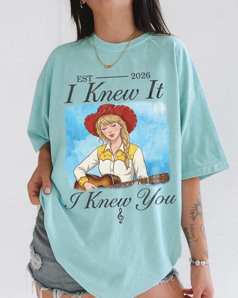 May include: A light blue t-shirt featuring a graphic of a person with a blonde braid wearing a red cowboy hat and a yellow and white shirt while playing an acoustic guitar. The text reads EST 2026 I Knew It I Knew You with a small musical note symbol.