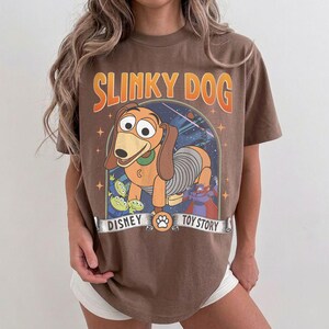 May include: Brown t-shirt with a graphic of Slinky Dog from Disney's Toy Story. The design includes Slinky Dog, the aliens, and the text "SLINKY DOG", "DISNEY", and "TOY STORY". The shirt has a vintage, distressed appearance.