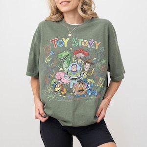 K&ouml;nnte beinhalten: Olivgr&uuml;nes kurz&auml;rmeliges T-Shirt mit einer farbenfrohen Grafik von Charakteren aus dem Animationsfilm "Toy Story". Das Design umfasst Woody, Buzz Lightyear, Rex und andere Charaktere. Die Worte "TOY STORY" sind oben auf der Grafik aufgedruckt.