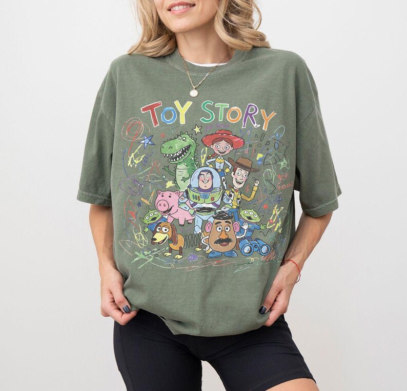 K&ouml;nnte beinhalten: Olivgr&uuml;nes kurz&auml;rmeliges T-Shirt mit einer farbenfrohen Grafik von Charakteren aus dem Animationsfilm "Toy Story". Das Design umfasst Woody, Buzz Lightyear, Rex und andere Charaktere. Die Worte "TOY STORY" sind oben auf der Grafik aufgedruckt.