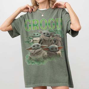 May include: An olive green short-sleeve t-shirt featuring a graphic print of multiple Grogu characters in tan robes surrounded by green smoke. The text GROGU is printed in large, bold, lime green letters above the characters on the front of the shirt.