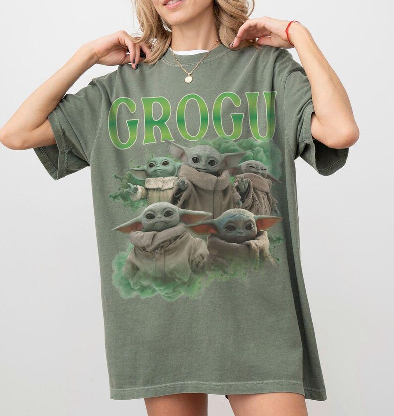 May include: An olive green short-sleeve t-shirt featuring a graphic print of multiple Grogu characters in tan robes surrounded by green smoke. The text GROGU is printed in large, bold, lime green letters above the characters on the front of the shirt.
