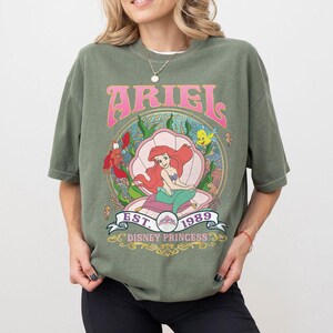 May include: An olive green t-shirt with a graphic of Ariel from The Little Mermaid. The design includes the word "ARIEL" in pink, the year "1989", and the words "Disney Princess". The graphic is set within a seashell.