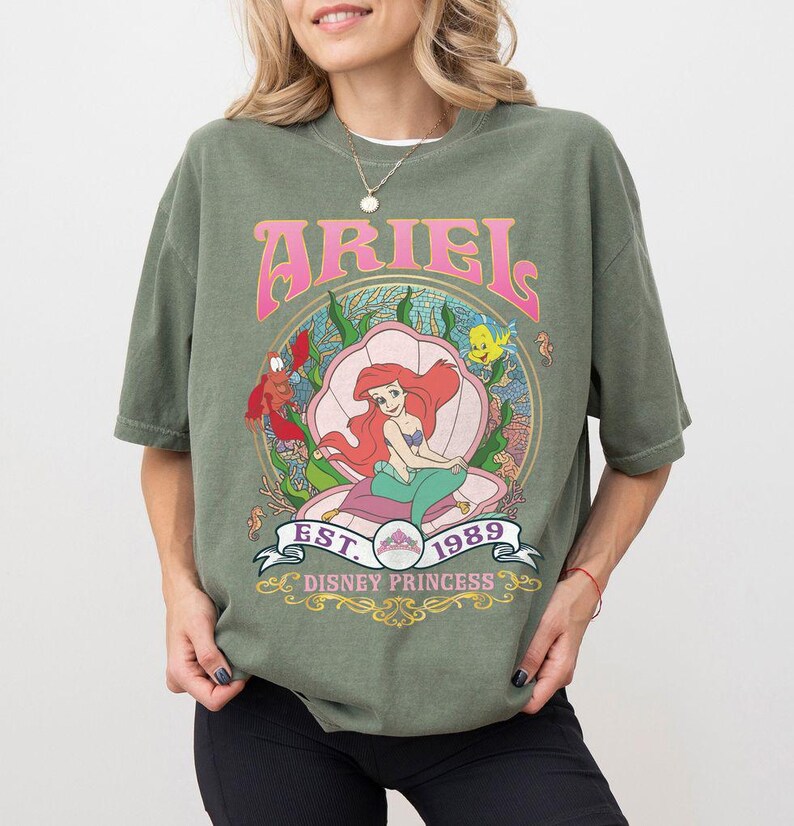 May include: An olive green t-shirt with a graphic of Ariel from The Little Mermaid. The design includes the word "ARIEL" in pink, the year "1989", and the words "Disney Princess". The graphic is set within a seashell.