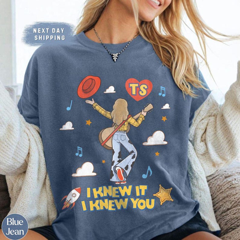 Puede incluir: Una camiseta azul con un gr&aacute;fico de una persona de pelo largo rubio que lleva una camisa de cuadros amarilla y pantalones con estampado de vaca, sosteniendo una guitarra. El dise&ntilde;o incluye un sombrero rojo, un coraz&oacute;n con el texto TS, nubes, estrellas, un cohete y notas musicales, con el texto I KNEW IT I KNEW YOU.