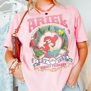 May include: Pale pink t-shirt with a graphic of Ariel from The Little Mermaid, inside a seashell, with the word "ARIEL" above and "EST. 1989 DISNEY PRINCESS" below. The design includes sea creatures and coral.