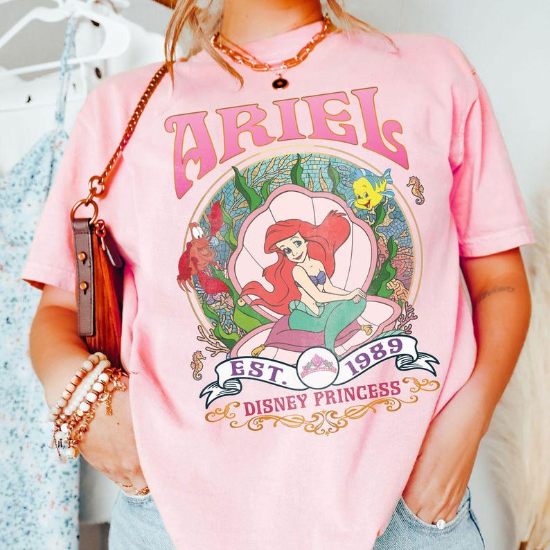 May include: Pale pink t-shirt with a graphic of Ariel from The Little Mermaid, inside a seashell, with the word "ARIEL" above and "EST. 1989 DISNEY PRINCESS" below. The design includes sea creatures and coral.