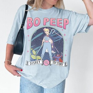 May include: Pale blue t-shirt with a graphic of Bo Peep from Toy Story. The design includes the text "BO PEEP" in pink, and the Disney and Toy Story logos. The shirt has short sleeves and a relaxed fit.