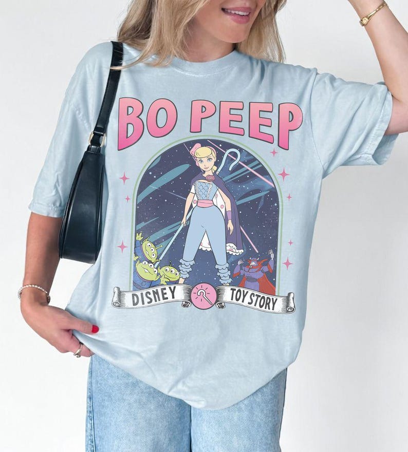 May include: Pale blue t-shirt with a graphic of Bo Peep from Toy Story. The design includes the text "BO PEEP" in pink, and the Disney and Toy Story logos. The shirt has short sleeves and a relaxed fit.