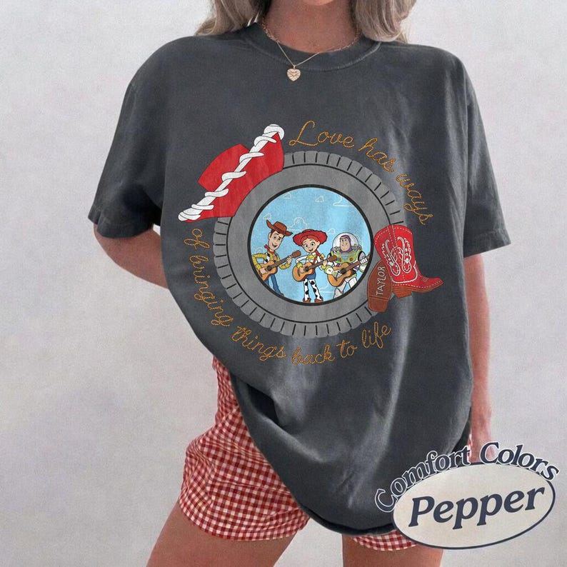 May include: A charcoal gray short-sleeve t-shirt featuring a circular graphic with Woody, Jessie, and Buzz Lightyear playing instruments. The design includes a red cowboy hat, a red boot labeled Taylor, and the text Love has ways of bringing things back to life.