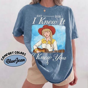 May include: A blue short-sleeve t-shirt featuring a graphic of a person with blonde hair wearing a red cowboy hat and a white shirt with yellow accents, playing an acoustic guitar. The text reads EST 2026 I Knew It I Knew You with a treble clef.