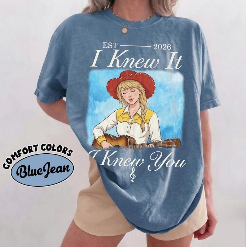 May include: A blue short-sleeve t-shirt featuring a graphic of a person with blonde hair wearing a red cowboy hat and a white shirt with yellow accents, playing an acoustic guitar. The text reads EST 2026 I Knew It I Knew You with a treble clef.