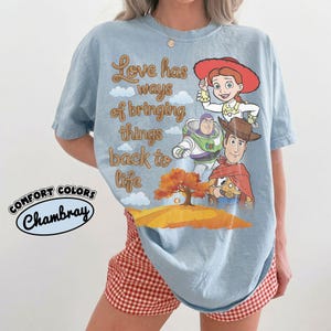 May include: A light blue t-shirt featuring a graphic print of Jessie, Buzz Lightyear, and Woody from Toy Story. The text reads Love has ways of bringing things back to life. Below the characters is an orange tree on a hill. Comfort Colors Chambray.