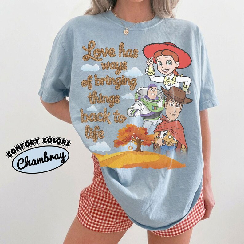 May include: A light blue t-shirt featuring a graphic print of Jessie, Buzz Lightyear, and Woody from Toy Story. The text reads Love has ways of bringing things back to life. Below the characters is an orange tree on a hill. Comfort Colors Chambray.