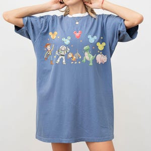 May include: A blue t-shirt featuring a graphic of Toy Story characters, including Woody, Buzz Lightyear, and Rex. The design includes Mickey Mouse-shaped balloons in various colors. The shirt has a white collar.