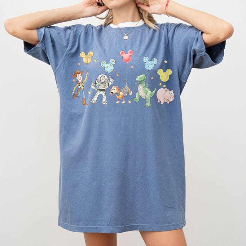 May include: A blue t-shirt featuring a graphic of Toy Story characters, including Woody, Buzz Lightyear, and Rex. The design includes Mickey Mouse-shaped balloons in various colors. The shirt has a white collar.