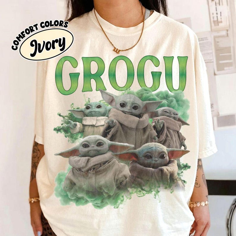 May include: An ivory t-shirt featuring the green text GROGU at the top and a graphic of four small, large-eared, green-skinned creatures surrounded by green smoke. A small oval label in the top left corner displays the text COMFORT COLORS IVORY.