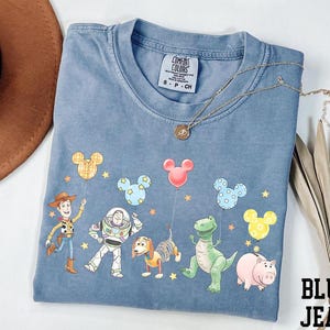 May include: Blue jean colored t-shirt featuring a graphic of Toy Story characters and Mickey Mouse balloons. The shirt has a crew neck and short sleeves. A gold necklace is draped across the shirt.