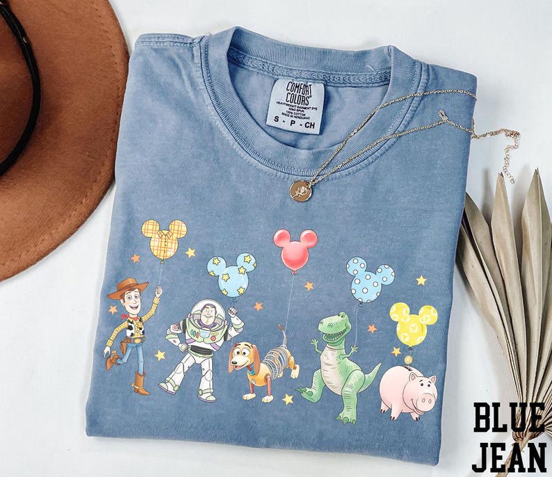 May include: Blue jean colored t-shirt featuring a graphic of Toy Story characters and Mickey Mouse balloons. The shirt has a crew neck and short sleeves. A gold necklace is draped across the shirt.