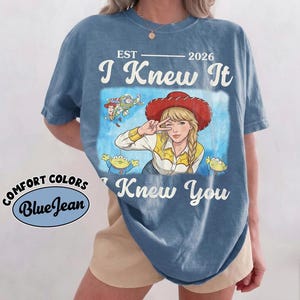 May include: A blue short-sleeve t-shirt featuring a graphic print of a person wearing a red cowboy hat, surrounded by cartoon characters. The text reads EST 2026 I Knew It I Knew You. A circular logo on the side displays the text COMFORT COLORS BlueJean.