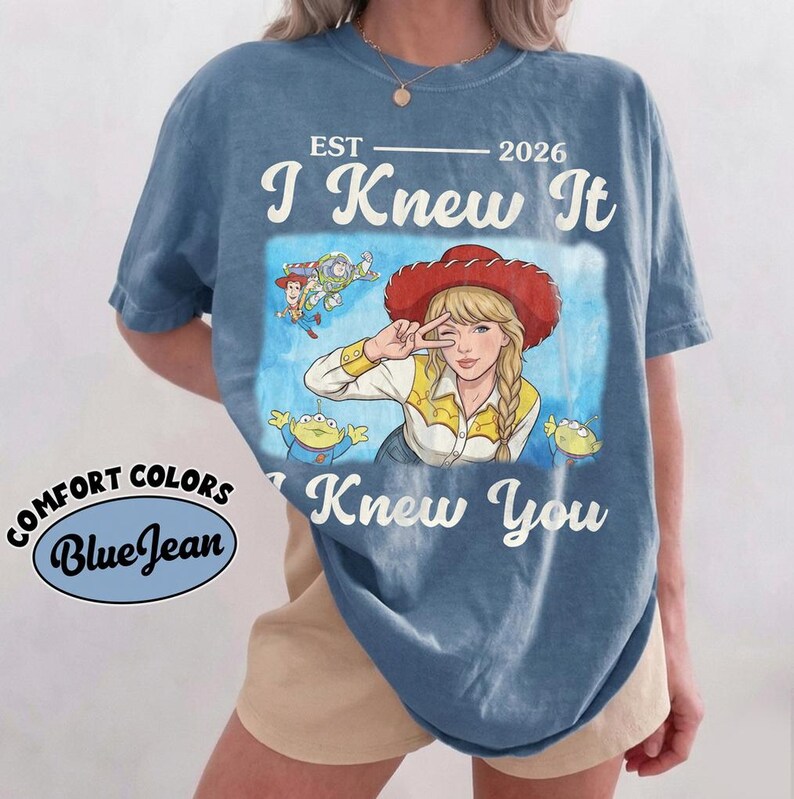 May include: A blue short-sleeve t-shirt featuring a graphic print of a person wearing a red cowboy hat, surrounded by cartoon characters. The text reads EST 2026 I Knew It I Knew You. A circular logo on the side displays the text COMFORT COLORS BlueJean.
