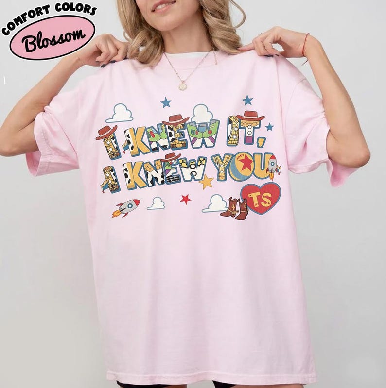 May include: A light pink short-sleeve t-shirt featuring a graphic print with the text I KNEW IT, I KNEW YOU. The design includes colorful cartoon elements like cowboy hats, rockets, clouds, stars, and a red heart with the letters TS inside.