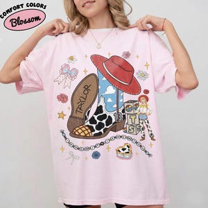 May include: A light pink t-shirt featuring a graphic print of a cowboy boot with a cow print pattern, a red hat, a character, and blocks with the letters T, S, and 13. The text "TAYLOR" and "I KNEW IT Y I KNEW Y OU" are printed on the shirt.